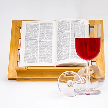 Ein offenes Buch auf einem Bambus-Leseständer. Davor stehen ein volles Glas Rotwein und ein umgekipptes leeres Weinglas. Eine sorgfältig arrangierte Szene mit einem geöffneten Buch auf einem Bambus-Leseständer. Ein volles Rotweinglas steht prominent im Vordergrund, flankiert von einem liegenden, leeren Weinglas, was eine Geschichte von Genuss und Ruhe erzählt.